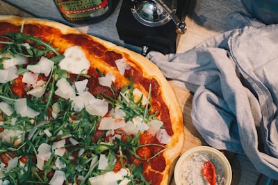 A freshly baked pizza topped with arugula, slices of parmesan cheese, and tomato sauce is placed on a wooden surface. Beside the pizza, there is a dark cloth napkin, a small bowl of coarse salt with a red spoon, and a can of olive oil. The pizza has a crispy crust and vibrant toppings that give it an appetizing look.