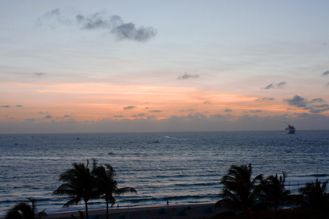 A vibrant sunset over a tropical beach with palm trees gently swaying and a cruise ship on the horizon.