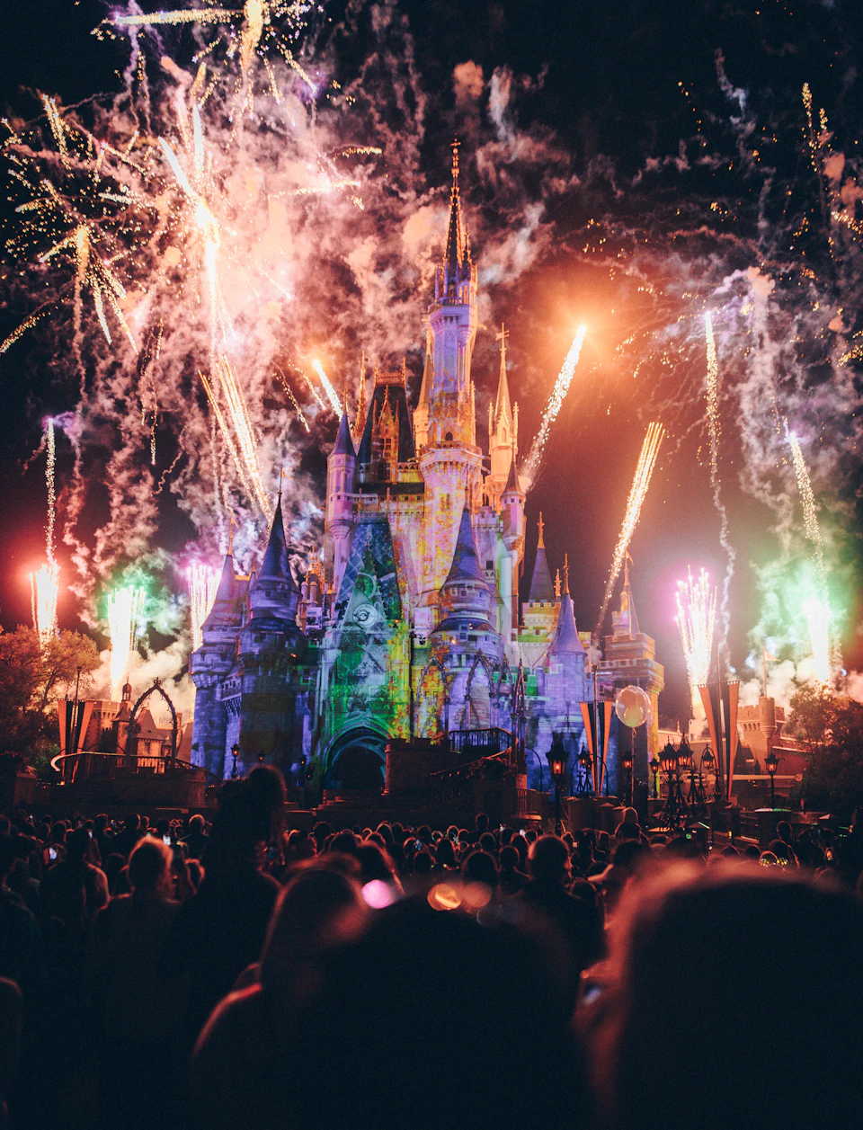 Crowd gathers around illuminated castle-like structure with fireworks exploding in the night sky