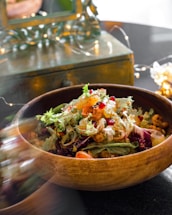 A wooden bowl filled with a colorful salad containing a mix of leafy greens, vibrant citrus slices, pomegranate seeds, and grilled chicken. Softly lit fairy lights and a reflective surface in the background create a warm, inviting atmosphere.