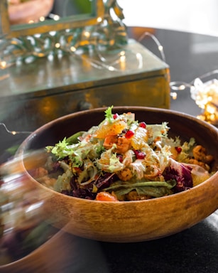 A wooden bowl filled with a colorful salad containing a mix of leafy greens, vibrant citrus slices, pomegranate seeds, and grilled chicken. Softly lit fairy lights and a reflective surface in the background create a warm, inviting atmosphere.