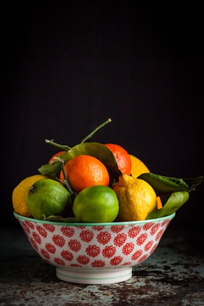 A decorative bowl with red floral patterns contains a variety of citrus fruits, including oranges, lemons, and limes, along with some green leaves. The setting is dimly lit, creating a focus on the vibrant colors of the fruits against the dark background.