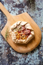 A rustic galette with a golden, flaky crust topped with slices of heirloom tomatoes and herbs sits on a wooden cutting board. The background features a distressed, textured surface in shades of blue and brown, adding an artisanal touch to the composition.