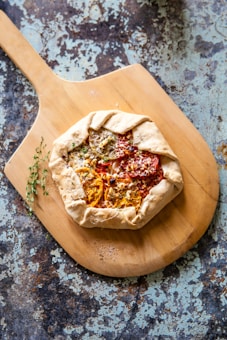 A rustic galette with a golden, flaky crust topped with slices of heirloom tomatoes and herbs sits on a wooden cutting board. The background features a distressed, textured surface in shades of blue and brown, adding an artisanal touch to the composition.