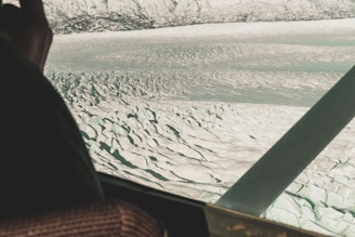 An aerial view of a vast, rugged glacier with deep crevasses and ridges. Snow-capped mountains are visible in the background, spanning across the horizon. A part of the airplane's window frame and interior, including a seat and armrest, are visible in the foreground.