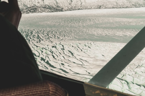 An aerial view of a vast, rugged glacier with deep crevasses and ridges. Snow-capped mountains are visible in the background, spanning across the horizon. A part of the airplane's window frame and interior, including a seat and armrest, are visible in the foreground.