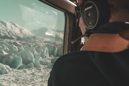 A person wearing a headset sits inside an aircraft, looking out at a vast expanse of jagged ice formations and distant snow-covered mountains. The interior of the aircraft is partially visible, enhancing the perspective of an aerial view.