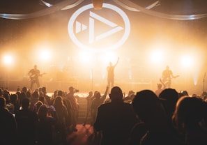 A live concert scene with a band performing on stage, illuminated by bright stage lights. A large crowd of people, some holding up phones, are gathered in front of the stage. The stage backdrop features a prominent logo and design, contributing to the energetic atmosphere.