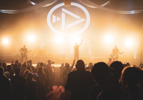 A live concert scene with a band performing on stage, illuminated by bright stage lights. A large crowd of people, some holding up phones, are gathered in front of the stage. The stage backdrop features a prominent logo and design, contributing to the energetic atmosphere.