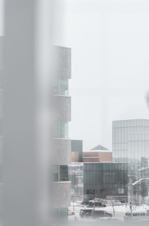 A cityscape featuring modern buildings with a minimalist design. The foreground reveals part of a structure with glass windows, creating a layered and angular visual effect. Sparse trees, cars, and a snowy urban environment are visible.