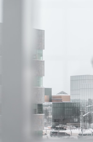 A cityscape featuring modern buildings with a minimalist design. The foreground reveals part of a structure with glass windows, creating a layered and angular visual effect. Sparse trees, cars, and a snowy urban environment are visible.