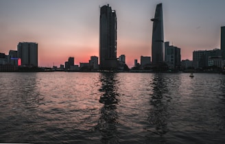 A city skyline at sunset with modern buildings and a calm river reflecting the lights.