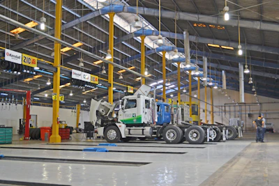 A large indoor vehicle maintenance facility with high ceilings and rows of overhead lights. Several trucks are parked in the space, with one having its hood open. Yellow support beams and signs hang from the ceiling. A person stands to the side, presumably working or supervising.
