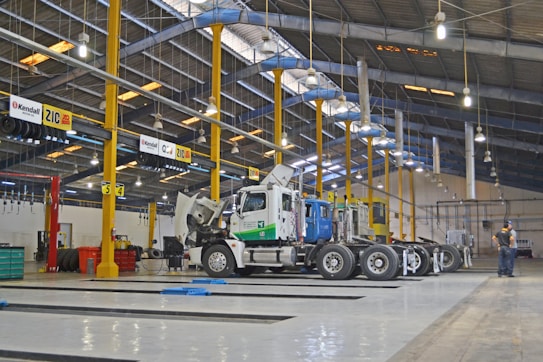 A large indoor vehicle maintenance facility with high ceilings and rows of overhead lights. Several trucks are parked in the space, with one having its hood open. Yellow support beams and signs hang from the ceiling. A person stands to the side, presumably working or supervising.