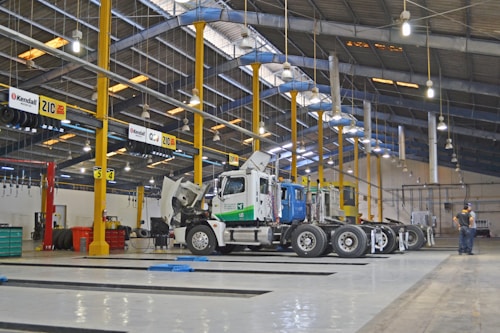 A large indoor vehicle maintenance facility with high ceilings and rows of overhead lights. Several trucks are parked in the space, with one having its hood open. Yellow support beams and signs hang from the ceiling. A person stands to the side, presumably working or supervising.