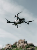 A drone hovers in the sky with a backdrop of rocky terrain. The quadcopter has a sleek, modern design with prominent propellers and a camera mounted at the front. The sky is partially cloudy, creating a serene and airy atmosphere.