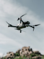 A drone hovers in the sky with a backdrop of rocky terrain. The quadcopter has a sleek, modern design with prominent propellers and a camera mounted at the front. The sky is partially cloudy, creating a serene and airy atmosphere.