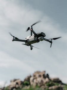 A drone hovers in the sky with a backdrop of rocky terrain. The quadcopter has a sleek, modern design with prominent propellers and a camera mounted at the front. The sky is partially cloudy, creating a serene and airy atmosphere.