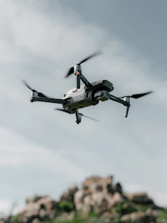 A drone hovers in the sky with a backdrop of rocky terrain. The quadcopter has a sleek, modern design with prominent propellers and a camera mounted at the front. The sky is partially cloudy, creating a serene and airy atmosphere.