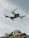 A drone hovers in the sky with a backdrop of rocky terrain. The quadcopter has a sleek, modern design with prominent propellers and a camera mounted at the front. The sky is partially cloudy, creating a serene and airy atmosphere.