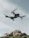 A drone hovers in the sky with a backdrop of rocky terrain. The quadcopter has a sleek, modern design with prominent propellers and a camera mounted at the front. The sky is partially cloudy, creating a serene and airy atmosphere.