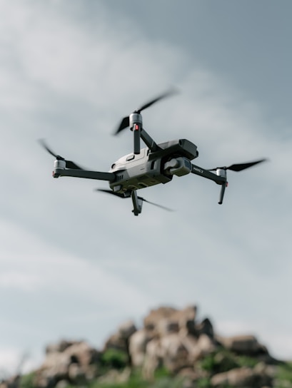 A drone hovers in the sky with a backdrop of rocky terrain. The quadcopter has a sleek, modern design with prominent propellers and a camera mounted at the front. The sky is partially cloudy, creating a serene and airy atmosphere.