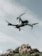 A drone hovers in the sky with a backdrop of rocky terrain. The quadcopter has a sleek, modern design with prominent propellers and a camera mounted at the front. The sky is partially cloudy, creating a serene and airy atmosphere.