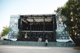 A large outdoor stage is set up with metal scaffolding and covered by a roof, surrounded by trees. The banners on either side read 'STRAND BÜHNE' and 'SINGOLDSAND FESTIVAL 2018'. The area in front of the stage appears empty, with some people walking and sitting nearby. Bright daylight illuminates the scene.