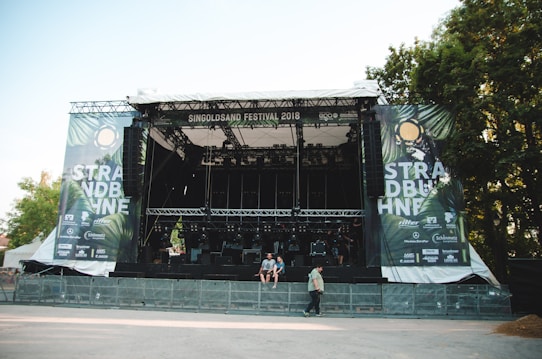 A large outdoor stage is set up with metal scaffolding and covered by a roof, surrounded by trees. The banners on either side read 'STRAND B&Uuml;HNE' and 'SINGOLDSAND FESTIVAL 2018'. The area in front of the stage appears empty, with some people walking and sitting nearby. Bright daylight illuminates the scene.