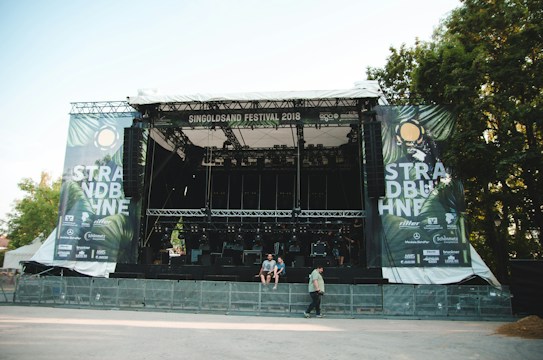 A large outdoor stage is set up with metal scaffolding and covered by a roof, surrounded by trees. The banners on either side read 'STRAND B&Uuml;HNE' and 'SINGOLDSAND FESTIVAL 2018'. The area in front of the stage appears empty, with some people walking and sitting nearby. Bright daylight illuminates the scene.