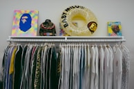 A clothing rack displaying a variety of white and colorful shirts, with a mix of plain and patterned designs. Positioned above the rack are several items including a colorful abstract artwork, a camouflage backpack, a yellow inflatable ring with text, and a book featuring a profile of a person.