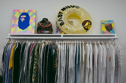 A clothing rack displaying a variety of white and colorful shirts, with a mix of plain and patterned designs. Positioned above the rack are several items including a colorful abstract artwork, a camouflage backpack, a yellow inflatable ring with text, and a book featuring a profile of a person.