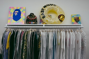 A clothing rack displaying a variety of white and colorful shirts, with a mix of plain and patterned designs. Positioned above the rack are several items including a colorful abstract artwork, a camouflage backpack, a yellow inflatable ring with text, and a book featuring a profile of a person.