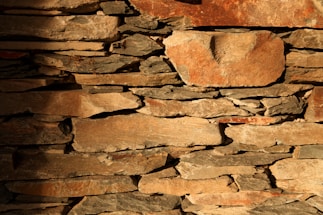 Roughly cut stones of various sizes are stacked irregularly to form a rustic wall. The stones are uneven in shape with a range of colors from light tan to darker brown hues. Some surfaces display weathering marks, creating a textured appearance with shadows enhancing the natural crevices between the stones.