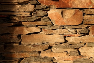 Roughly cut stones of various sizes are stacked irregularly to form a rustic wall. The stones are uneven in shape with a range of colors from light tan to darker brown hues. Some surfaces display weathering marks, creating a textured appearance with shadows enhancing the natural crevices between the stones.