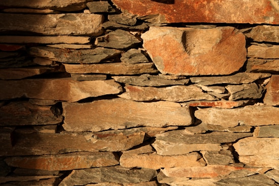 Roughly cut stones of various sizes are stacked irregularly to form a rustic wall. The stones are uneven in shape with a range of colors from light tan to darker brown hues. Some surfaces display weathering marks, creating a textured appearance with shadows enhancing the natural crevices between the stones.