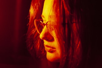 A person with long hair and glasses bathed in a deep red light, creating a warm and intense atmosphere.