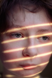 Soft sunlight filters through blinds, casting striped shadows across a pensive face. The gaze is directed slightly downward, creating a thoughtful and introspective mood.