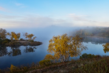A serene lakeside scene with mist hovering above the water. The landscape features vibrant autumn trees with golden leaves reflected in the calm lake. The distant shoreline fades into the mist, creating a peaceful and tranquil atmosphere.