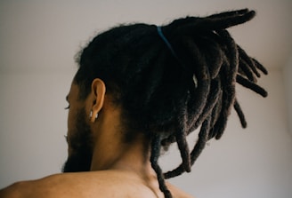 A person with their back turned is visible, showcasing their dreadlocked hair tied up with a blue band. The individual is wearing an earring in one ear, and the overall setting appears to be indoors with a neutral-colored wall in the background.