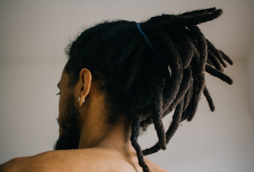 A person with their back turned is visible, showcasing their dreadlocked hair tied up with a blue band. The individual is wearing an earring in one ear, and the overall setting appears to be indoors with a neutral-colored wall in the background.