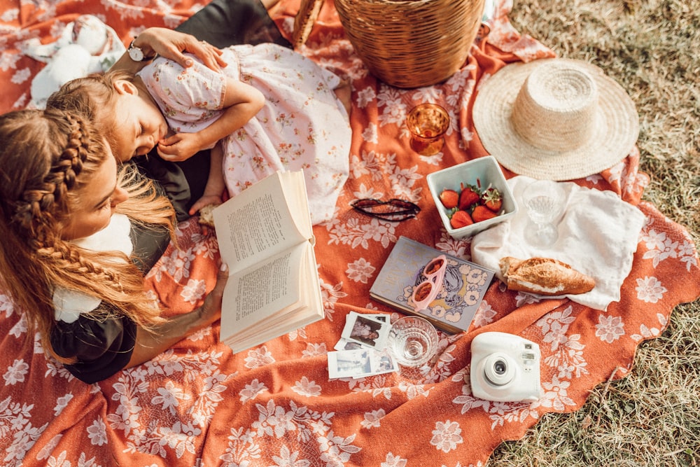 woman reading with girl while lying on orange and white floral picnic mat