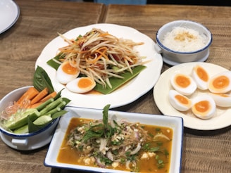 An assortment of dishes is displayed on a wooden table, featuring a central plate with a papaya salad accompanied by halved soft-boiled eggs. Surrounding it are sides including a bowl of fresh vegetable sticks, a plate of halved boiled eggs with orange yolks, a bowl of thin noodles with a sprinkle of garnish, and a rectangular dish of a mixed vegetable dish in a savory sauce garnished with mint leaves.