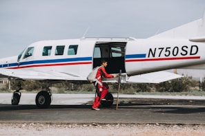 A person in a red jumpsuit is standing near a small airplane with an open door, preparing or inspecting equipment. The airplane is white with blue and red stripes and is parked on a tarmac with some vegetation in the background.