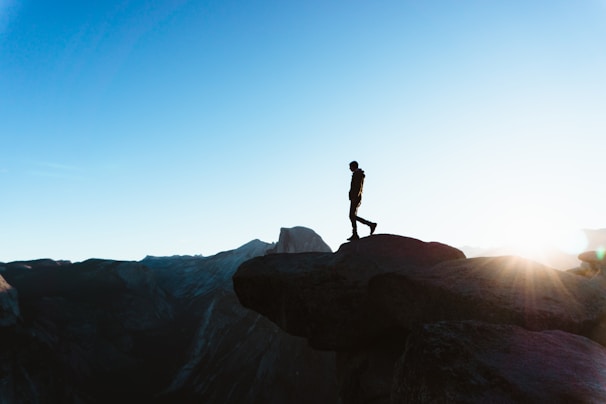 Sunset silhouette of a woman standing on a cliff overlooking a vast mountain range.
