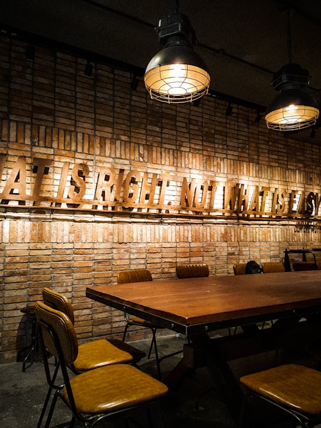 A rustic dining space featuring a long wooden table with brown leather chairs. The brick wall in the background is adorned with a prominent message made of metal letters. Several industrial-style pendant lights hang from the ceiling, casting warm light over the area.