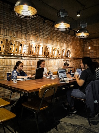 A group of people are working at a large wooden table in an industrial-style space with exposed brick walls. Overhead, industrial light fixtures provide warm lighting. Laptops and coffee cups are scattered across the table, indicating a collaborative or study environment. The phrase 'right, not what is easy' is prominently displayed on the wall in large letters.