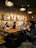 A group of people are working at a large wooden table in an industrial-style space with exposed brick walls. Overhead, industrial light fixtures provide warm lighting. Laptops and coffee cups are scattered across the table, indicating a collaborative or study environment. The phrase 'right, not what is easy' is prominently displayed on the wall in large letters.