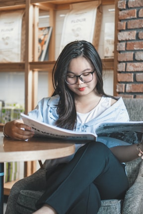 A person with long hair and glasses is reading a magazine while seated in a cozy room with wooden shelves and a brick wall. The setting appears to be comfortable and relaxed, with a focus on leisure and reading.