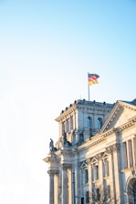 A historic architectural building with classical columns and statues. At the top, a German flag waves against a clear blue sky.